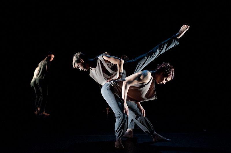 A man executes a penche while resting his hand on the small of another dancer's back